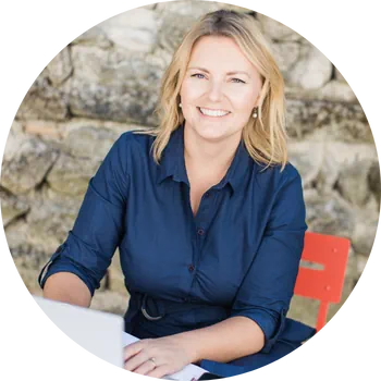 A woman in a blue shirt smiles while sitting at a table with her laptop, perhaps updating her WordPress maintenance plans, set against a stone wall backdrop.