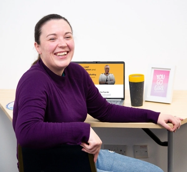 Kim Scotland, in a purple sweater, sits at a desk smiling with her laptop, contact mug, and a framed "You Go Girl" print in the background.